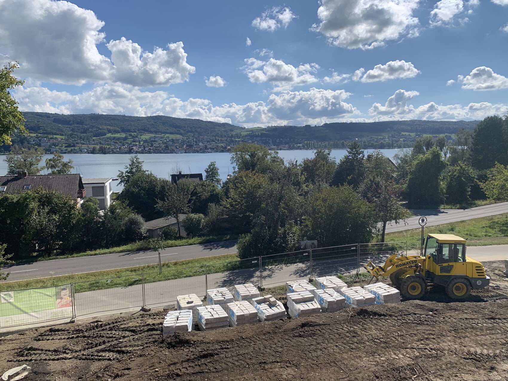 Construction site on a hillside: yellow front loader beside stacked pallets, view of lake and hills.