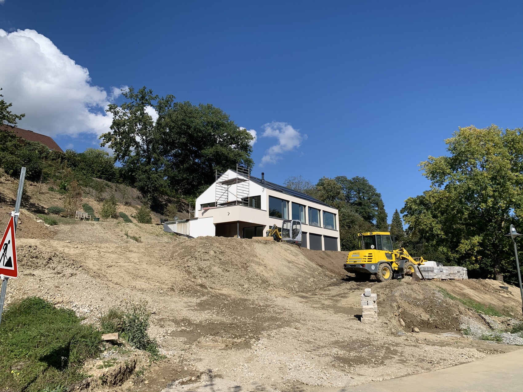 Modern white single-family house construction site on gravel mound; yellow excavator, scaffolding, trees, blue sky