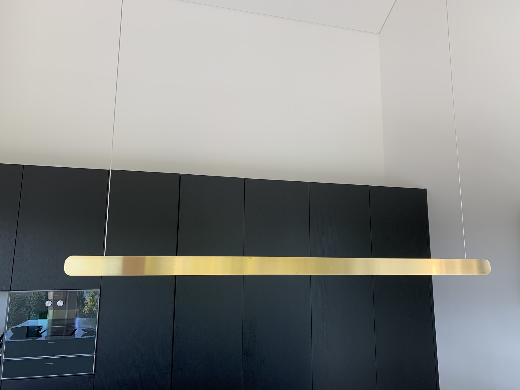 Long golden pendant fixture hangs horizontally above black kitchen cabinets in a bright room.