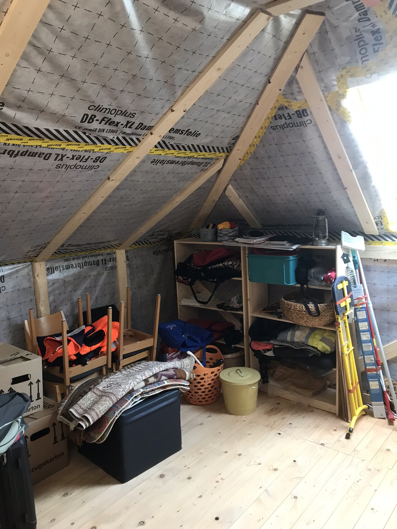 Attic with sloped roof, wooden beams, and insulation; shelves with clothes, boxes, and ladder.