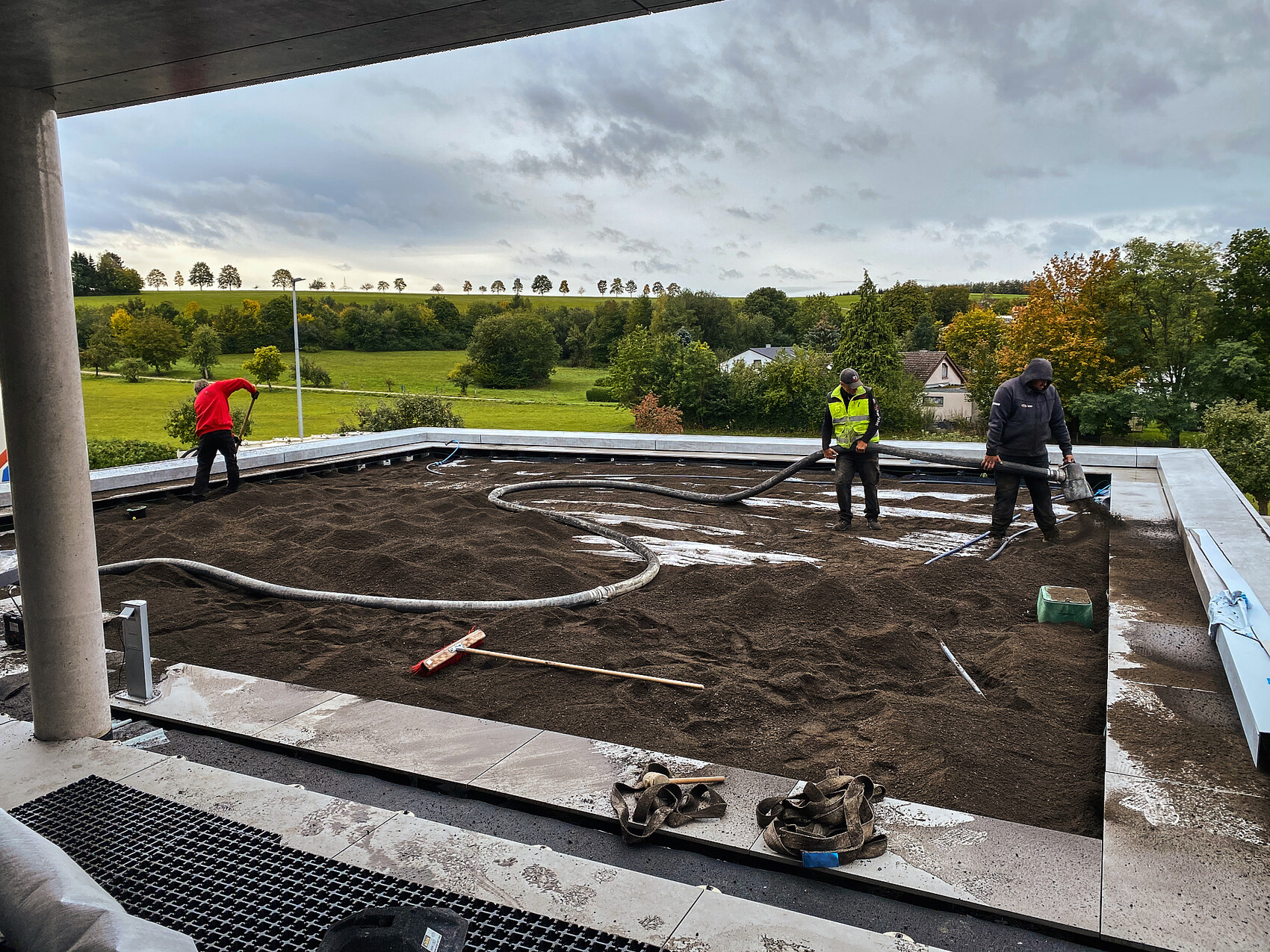 Three workers spreading soil on a flat roof; hose lying on it, landscape in the background.