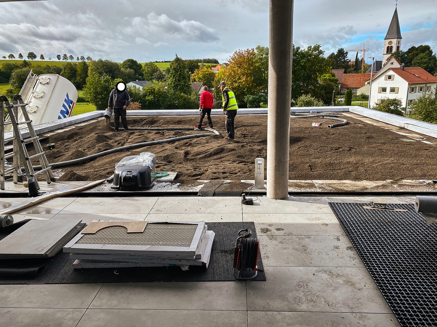 Construction workers spreading soil on flat roof; hoses, tools, village with church tower in the background.