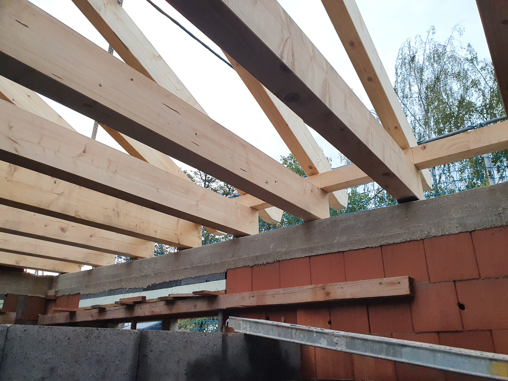 Wooden beam roof structure over a red brick wall; beams are laid crosswise, tree and sky in the background.