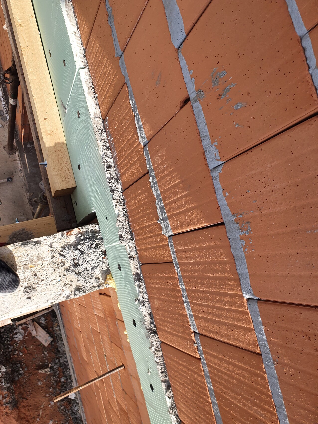Roof with red clay tiles, gray joints, green insulation material at the edge, and construction timber on site