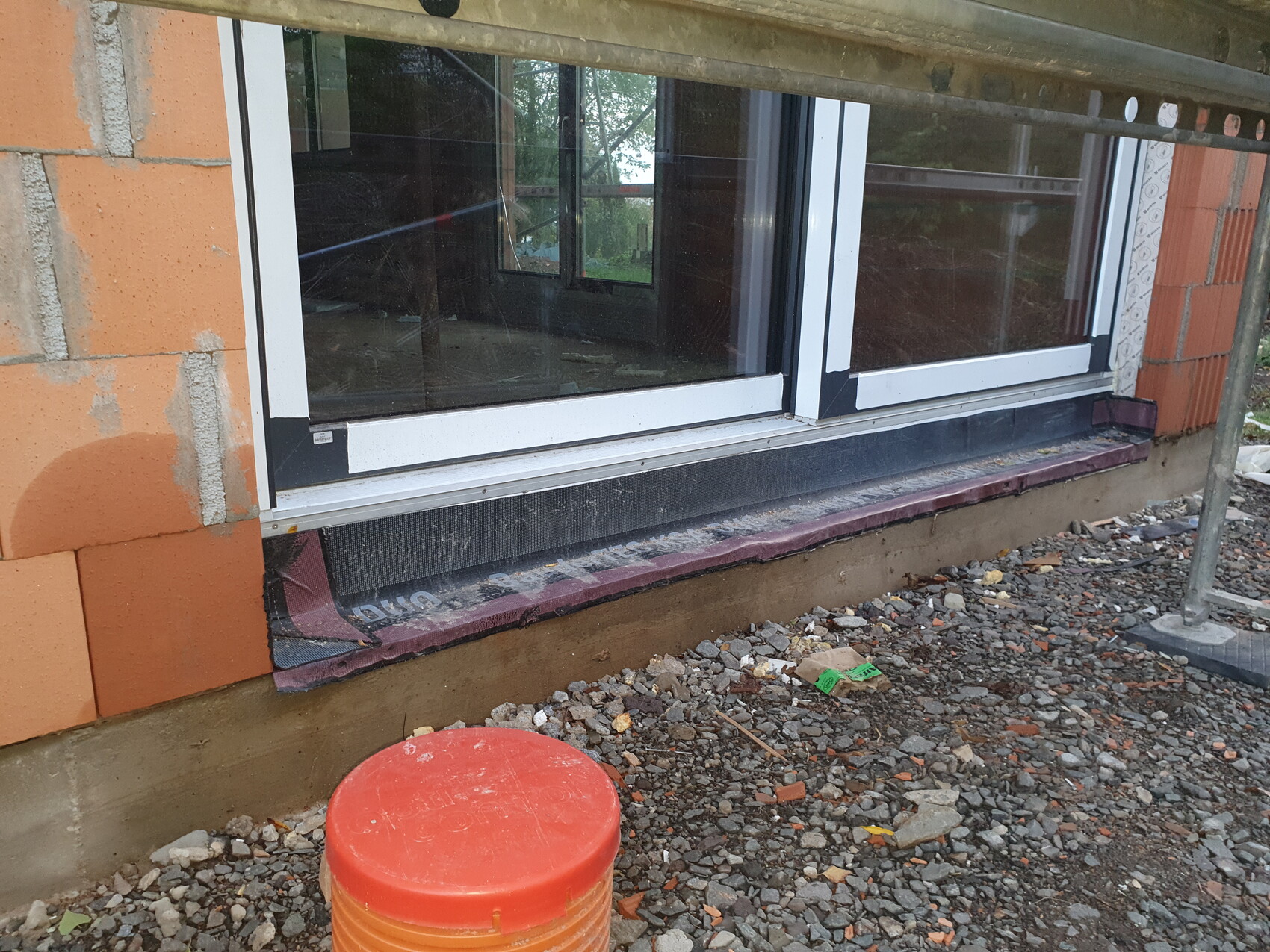 Construction site at an exterior wall with large window, white frame, black waterproof membrane; red bucket in front.