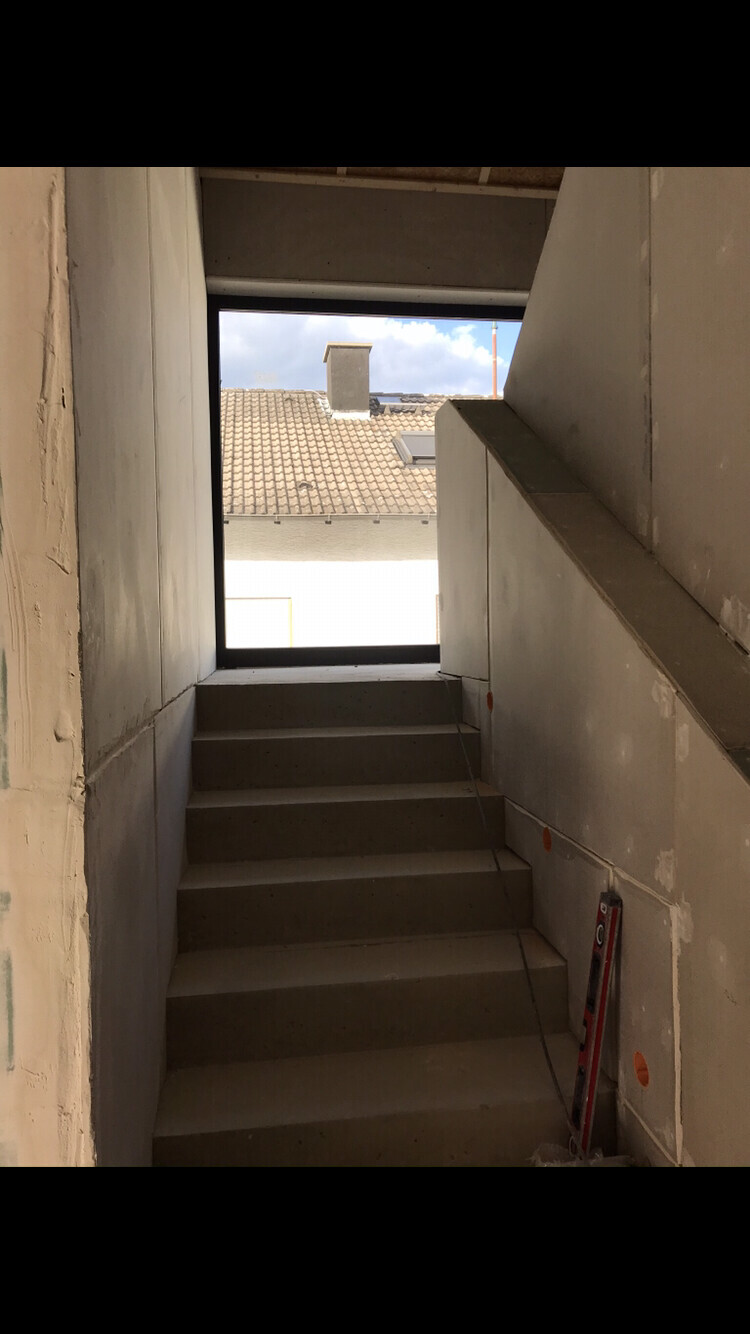 Unfinished concrete stairwell; view through upper opening to roof tiles and blue sky.