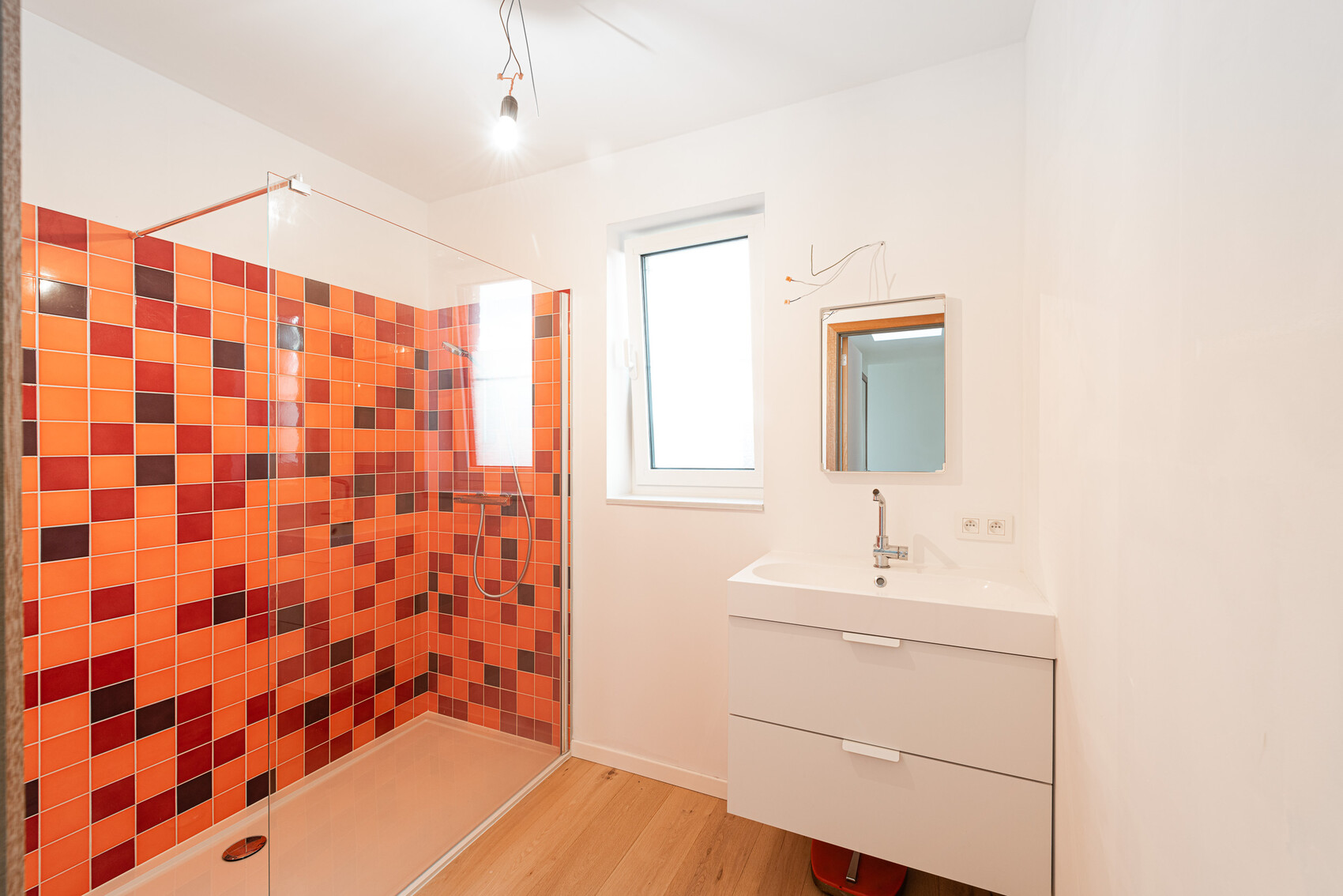 Bathroom with glass shower, orange-red tiles, white washbasin, window and mirror.