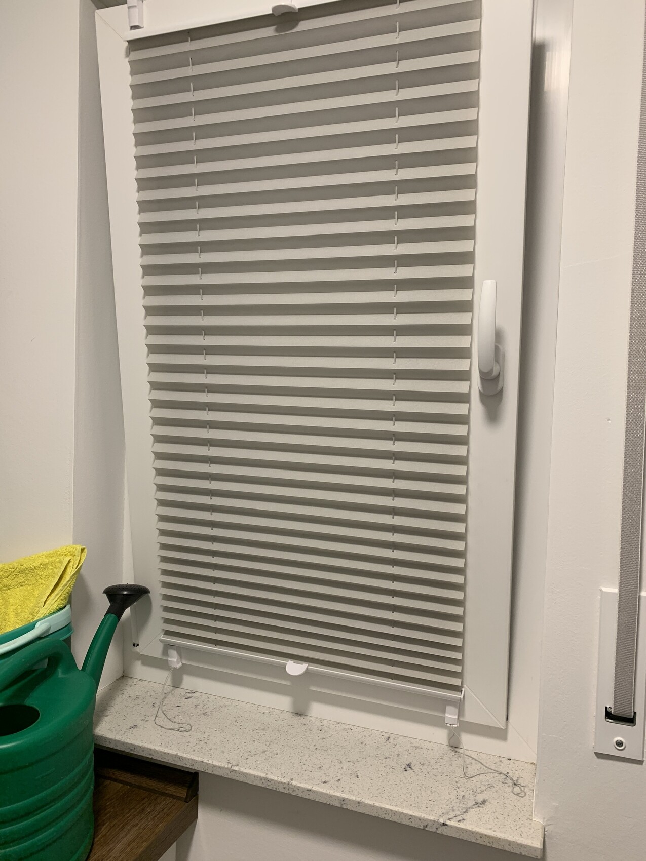 White window blind with closed slats; green watering can with yellow cloth next to windowsill.