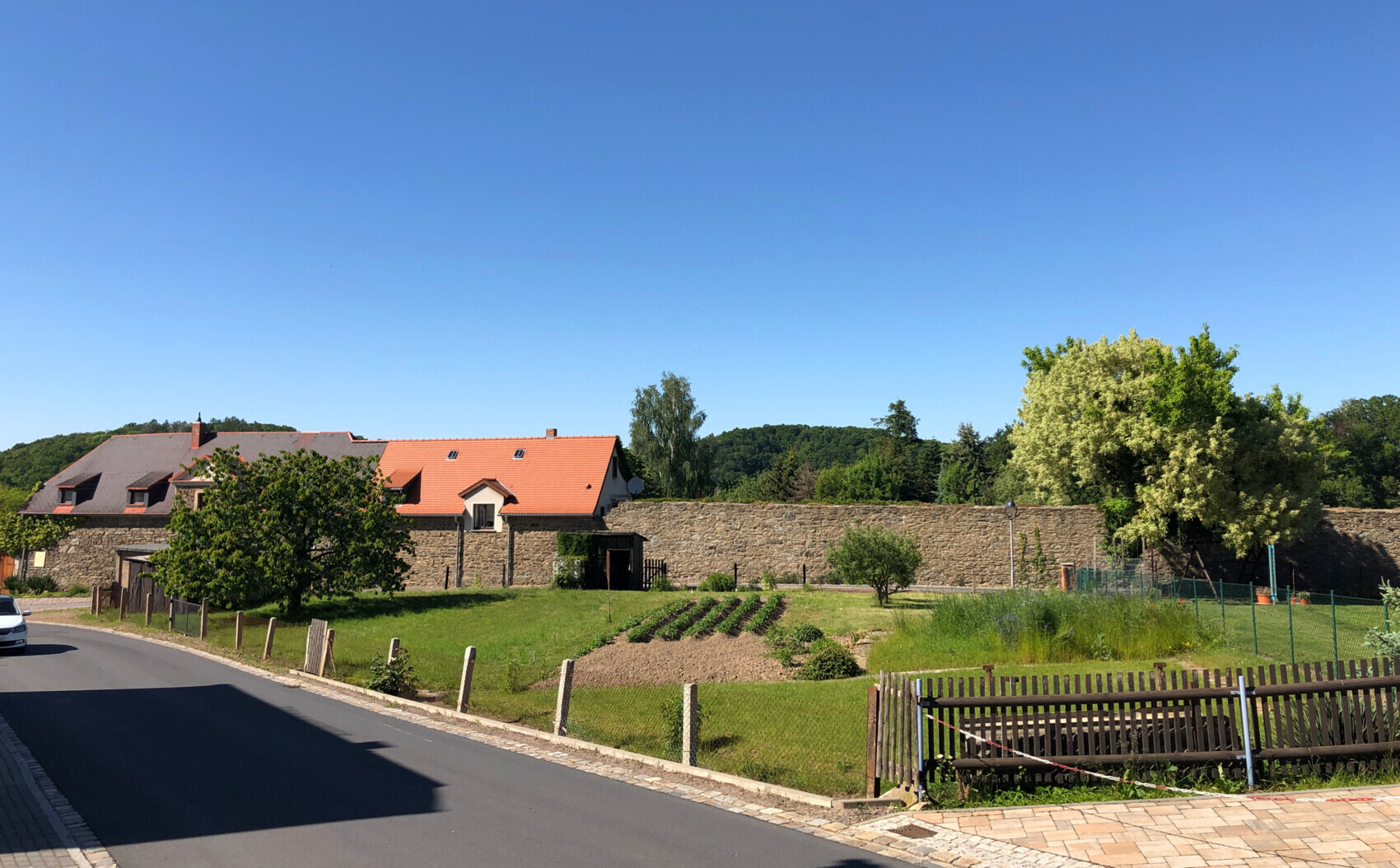 Rural scene: stone wall, houses with red tiled roofs, garden beds and trees in front of a green area.
