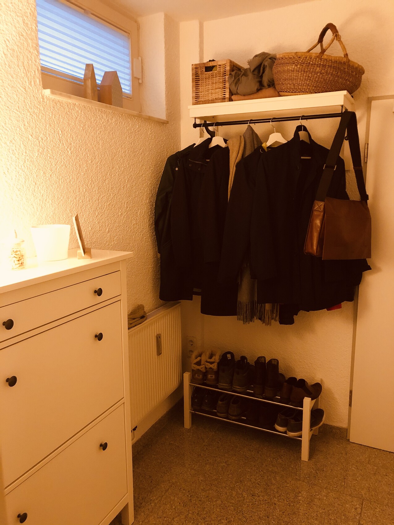 Wardrobe corner with coats on the rail, shoe rack below, baskets on top, white dresser on the left.