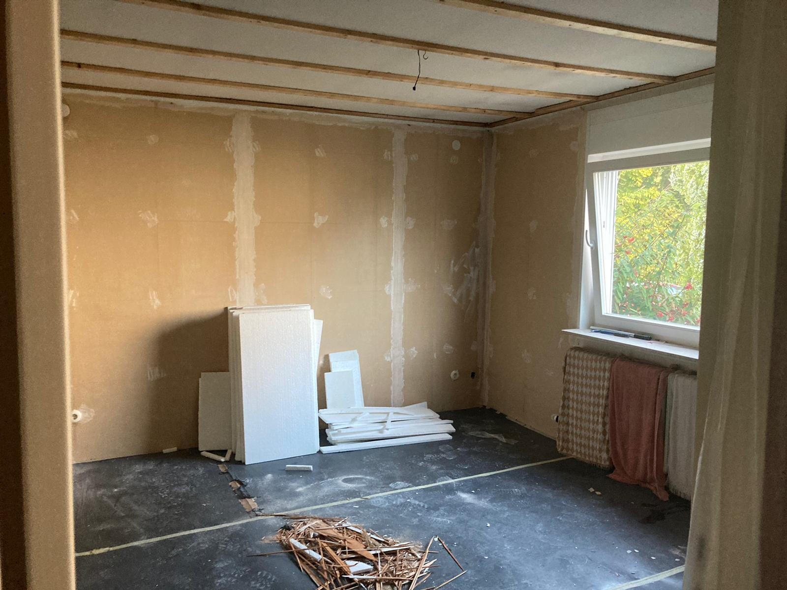 Renovierungsraum mit freigelegten Wänden, Bauplatten und Müll am Boden, Fenster sichtbar.