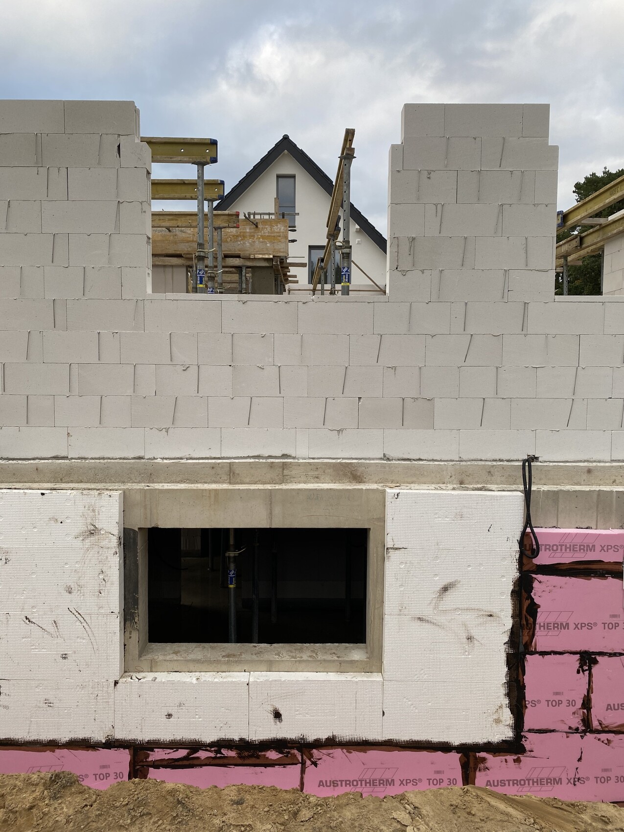 Shell construction view: white concrete blocks, foundation opening, and scaffolding in front of a house.