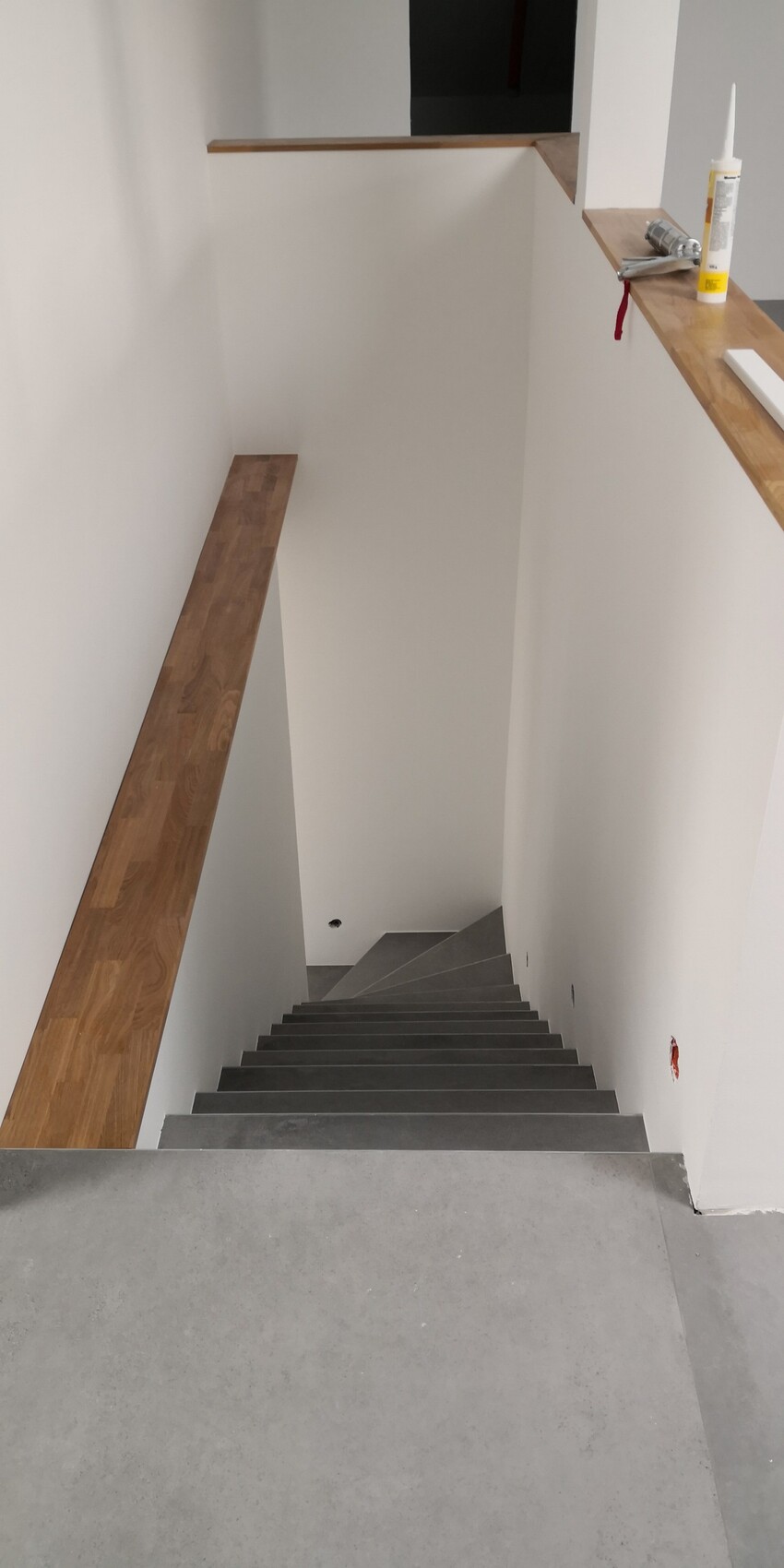 Interior view of an indoor staircase: gray steps, white walls, wooden handrail on the left.