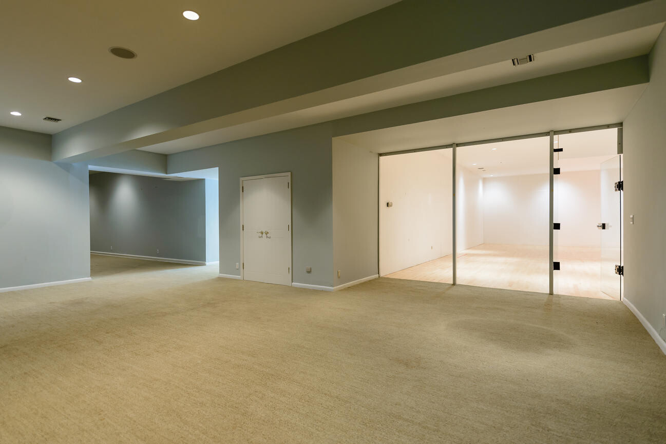 Large, empty basement room with bright walls; glass doors on the right leading to a bright room with wooden floor.