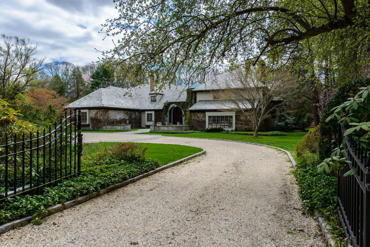 Small house with stone facade and curved gravel driveway, flanked by garden fences and trees.