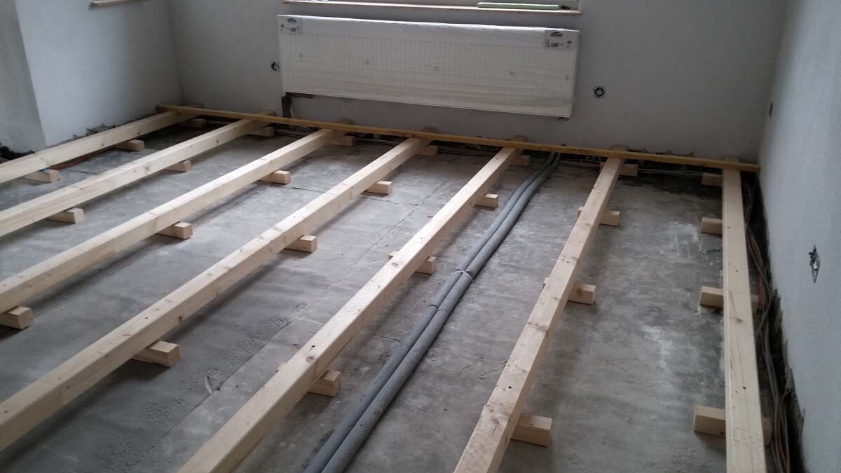 Room with wooden beam substructure on raw floor, radiator on the wall, cables on the floor.