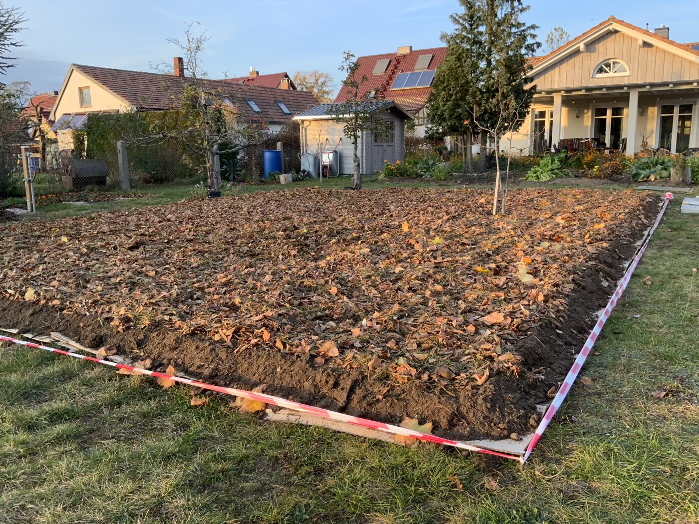 Rechteckige Gartenfläche mit Erdreich, Herbstlaub und Absperrband; Haus im Hintergrund.