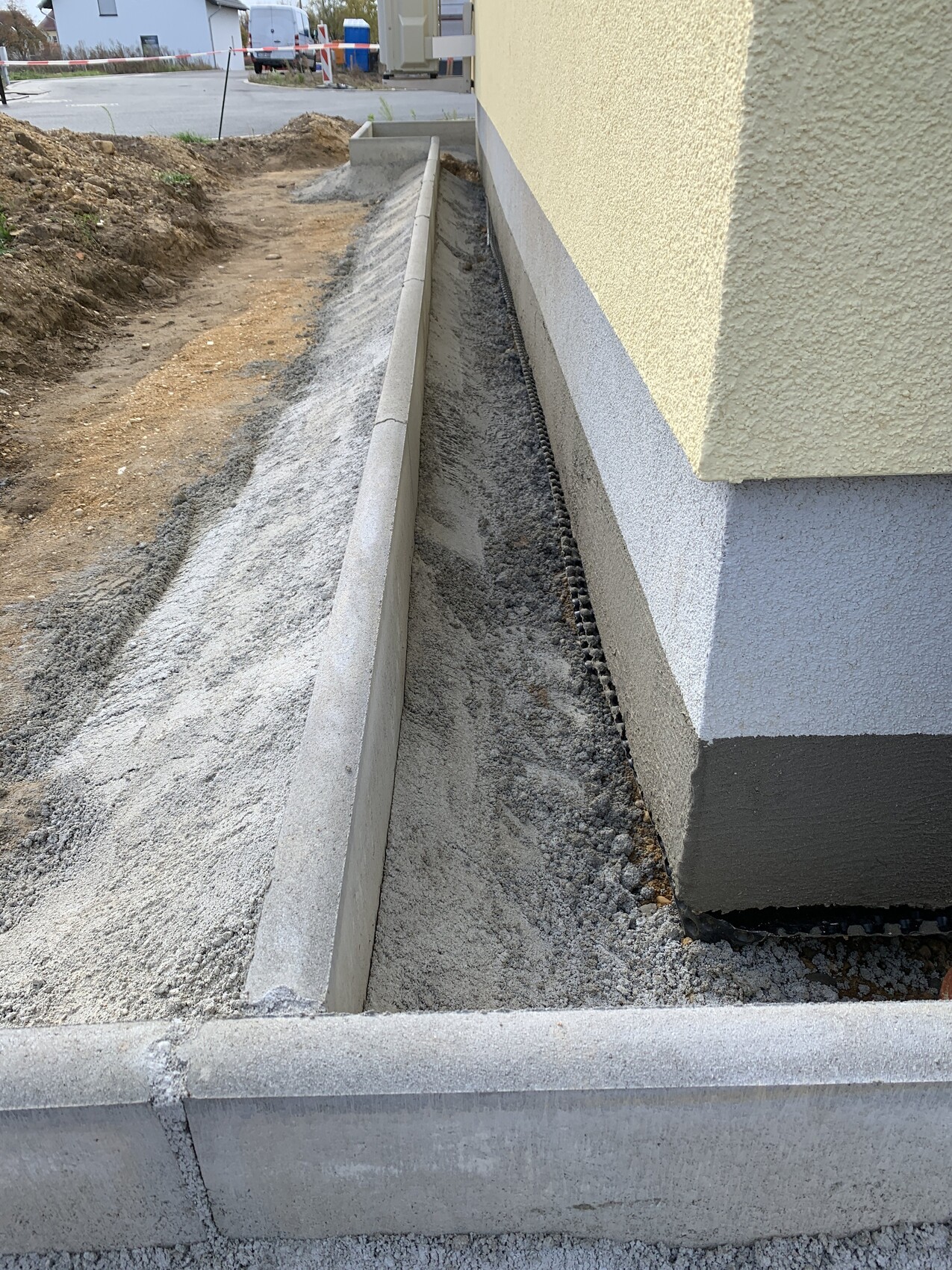 Construction site next to a yellow building; trench with gravel, concrete wall, barrier tape in the background.