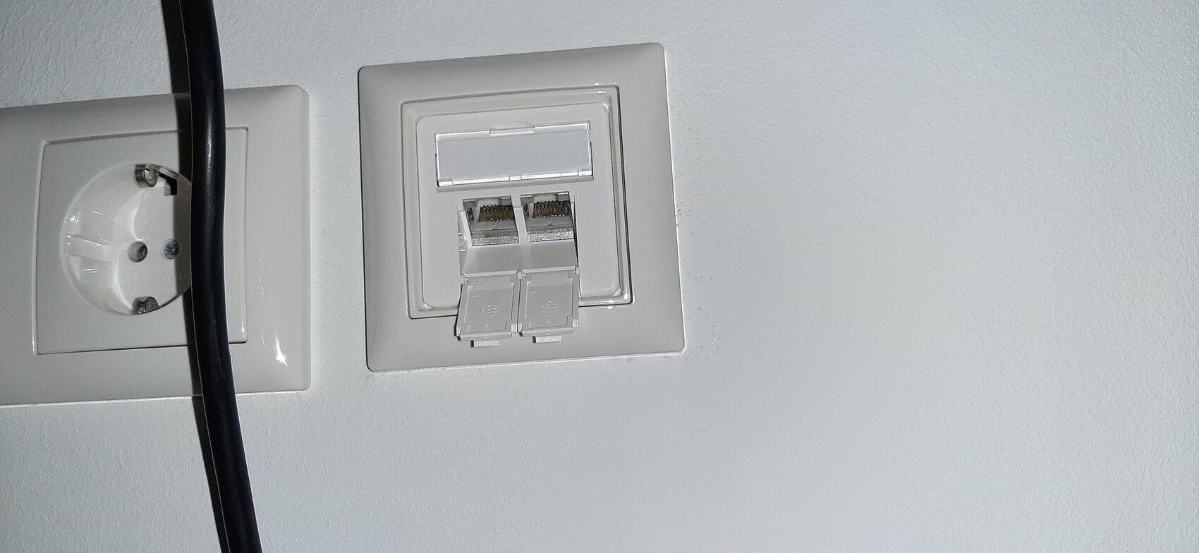 White wall plate with two RJ-45 network sockets and a round power outlet next to it; black cable in front.