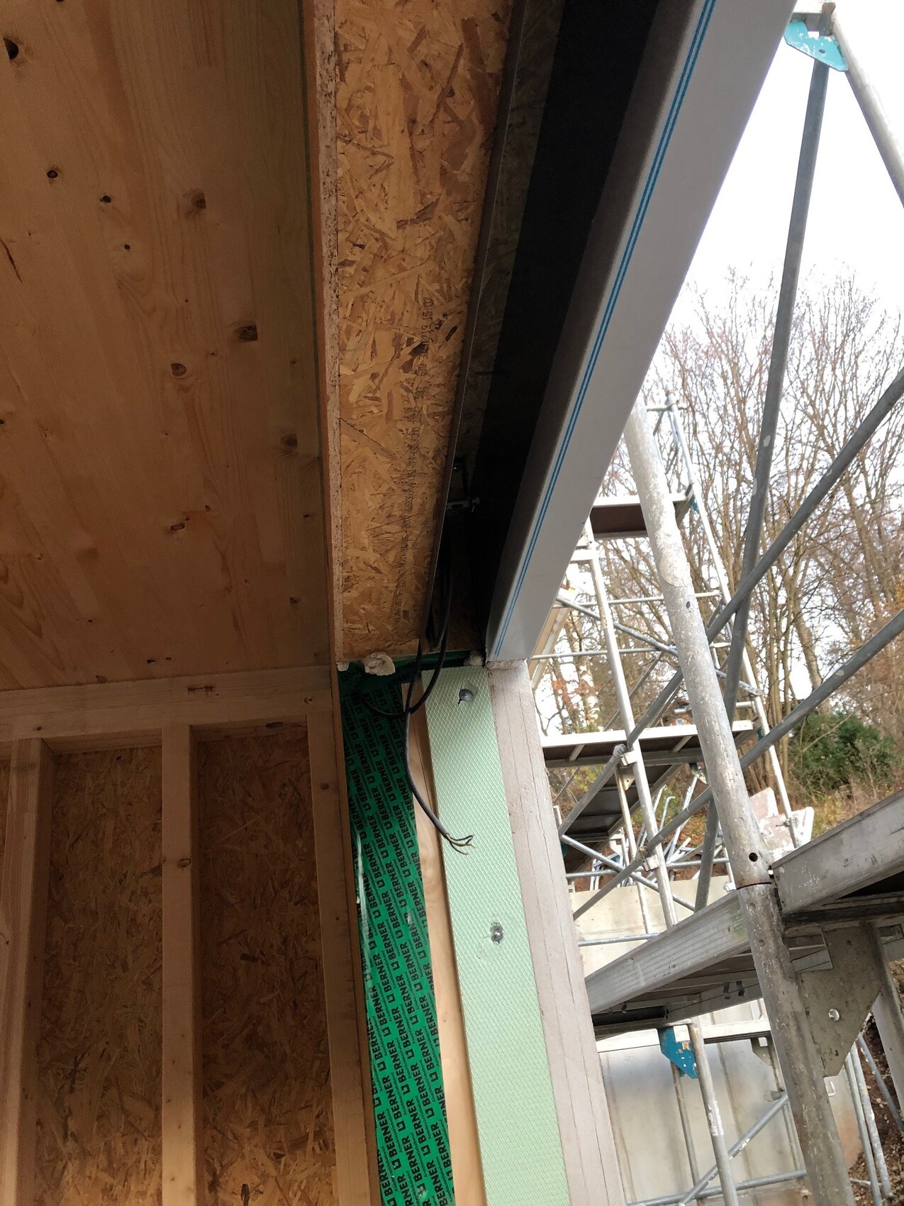 Construction site: timber frame construction with OSB boards, green insulation, scaffolding visible outside.