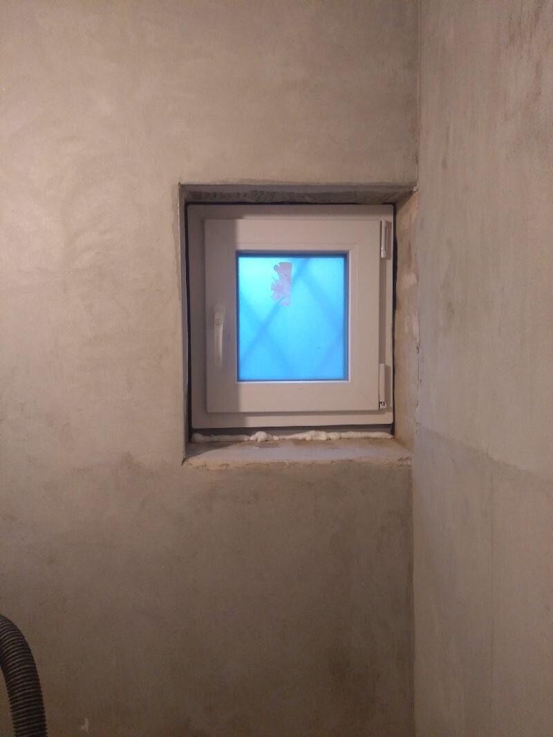 Small, square white window in untreated concrete wall; blue light behind glass.