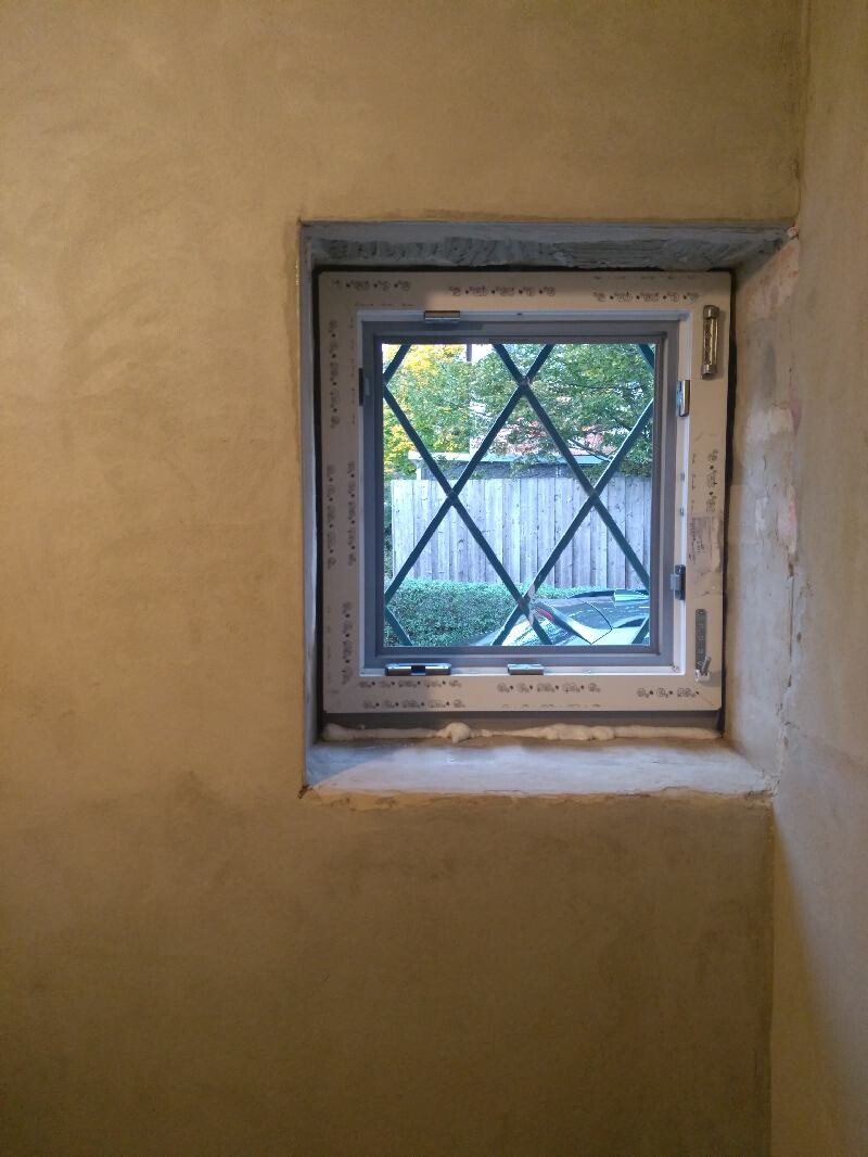 Window with metal cross grille in a rough interior wall; view outside to fence and garden.