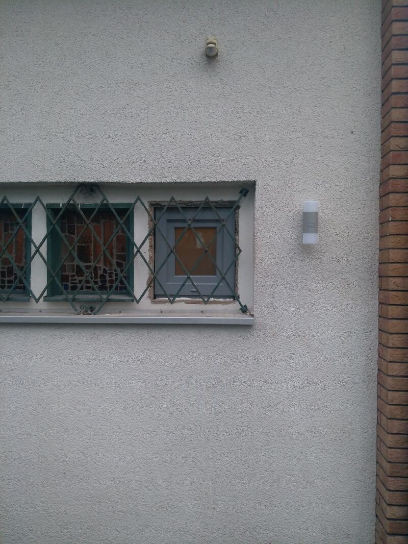 White wall with small window behind green metal grille and side exterior light.