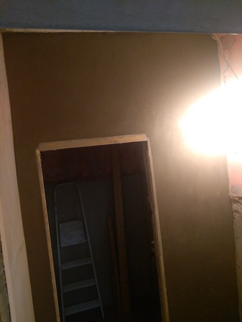 Construction basement: open door frame, ladder in a dark room, wooden slats; bright ceiling light on the right.