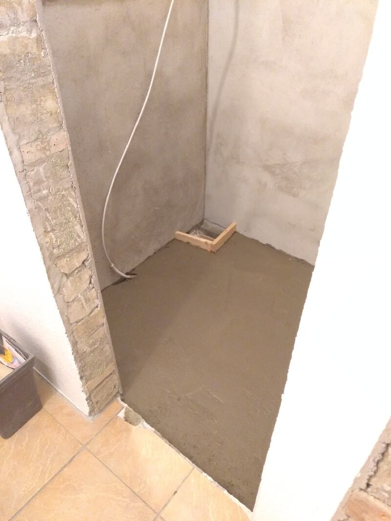 Wet concrete floor in a corner, left brick wall, cables on the wall, wooden frame around the drain.
