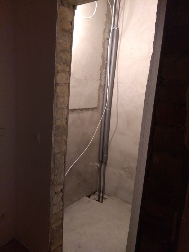 Unrenovated passage with exposed pipes, cables, and brick wall.