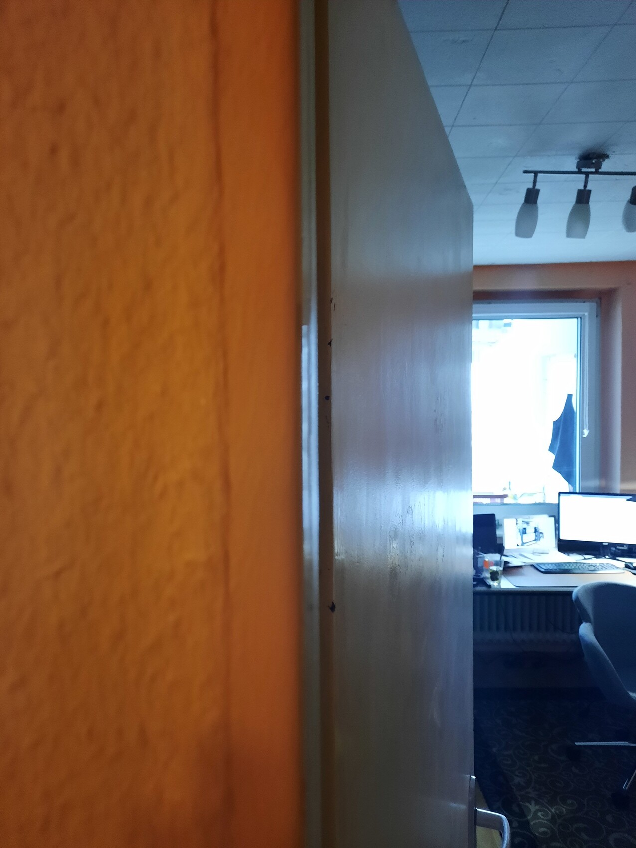View through a partly open door into an office with desk, monitors, chair, and window.
