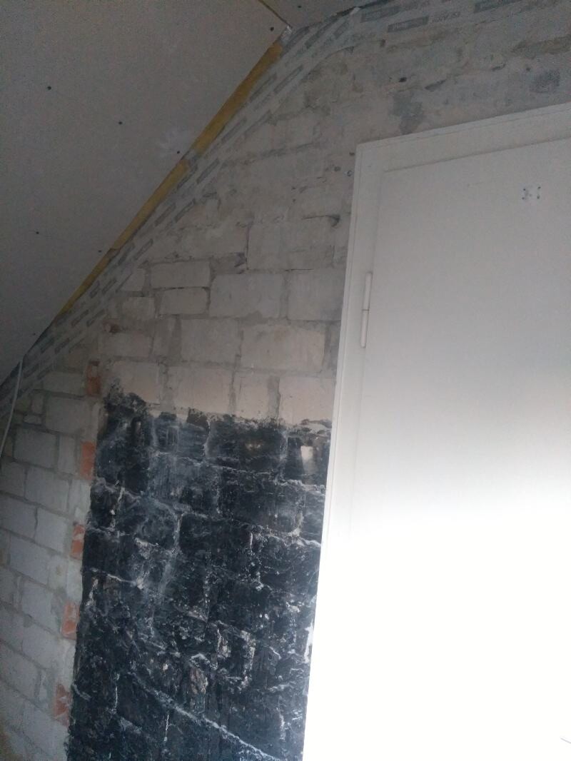 Unplastered brick wall, black area at the bottom, white door on the right, left sloping roof.