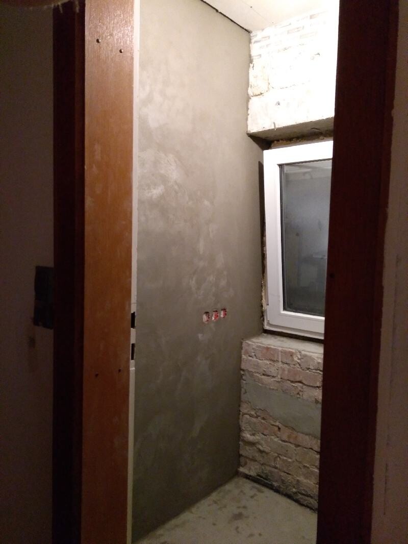 Basement corner room with gray plaster wall, exposed bricks under the window, and white frame.