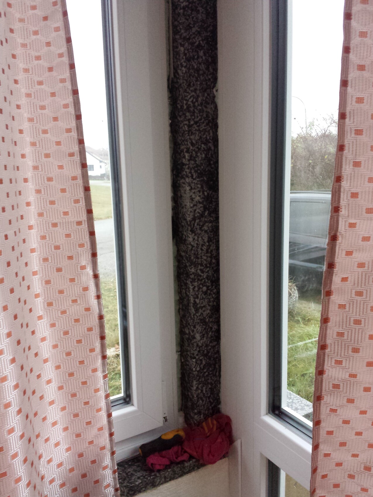 Window with pink curtains; between them a dark, textured column; fabric scraps on the windowsill.