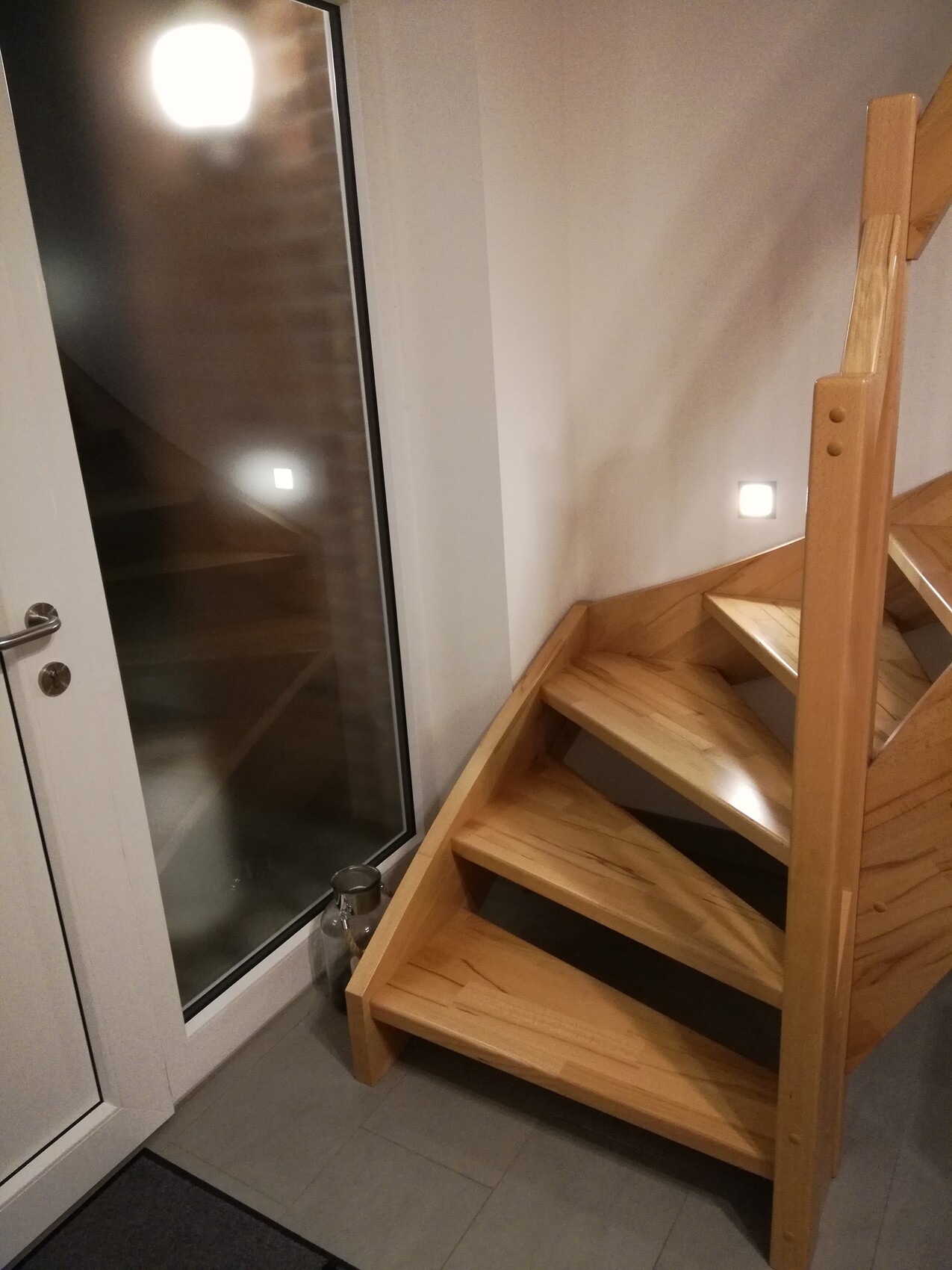 Holztreppe im Flur neben Glastür, helle Wände und kleine Wandleuchten.