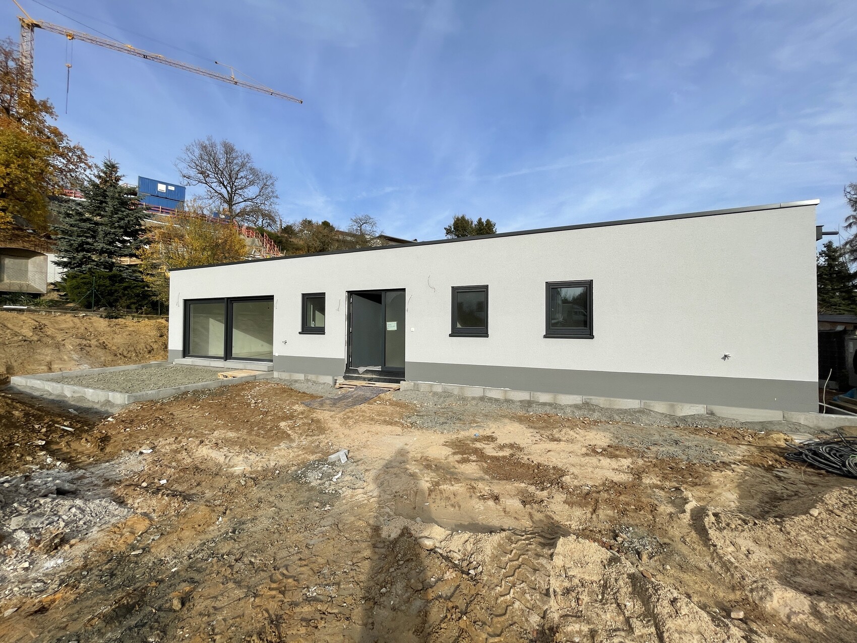 New construction single building with flat roof, white facade, windows/door, construction pit, crane in the background.
