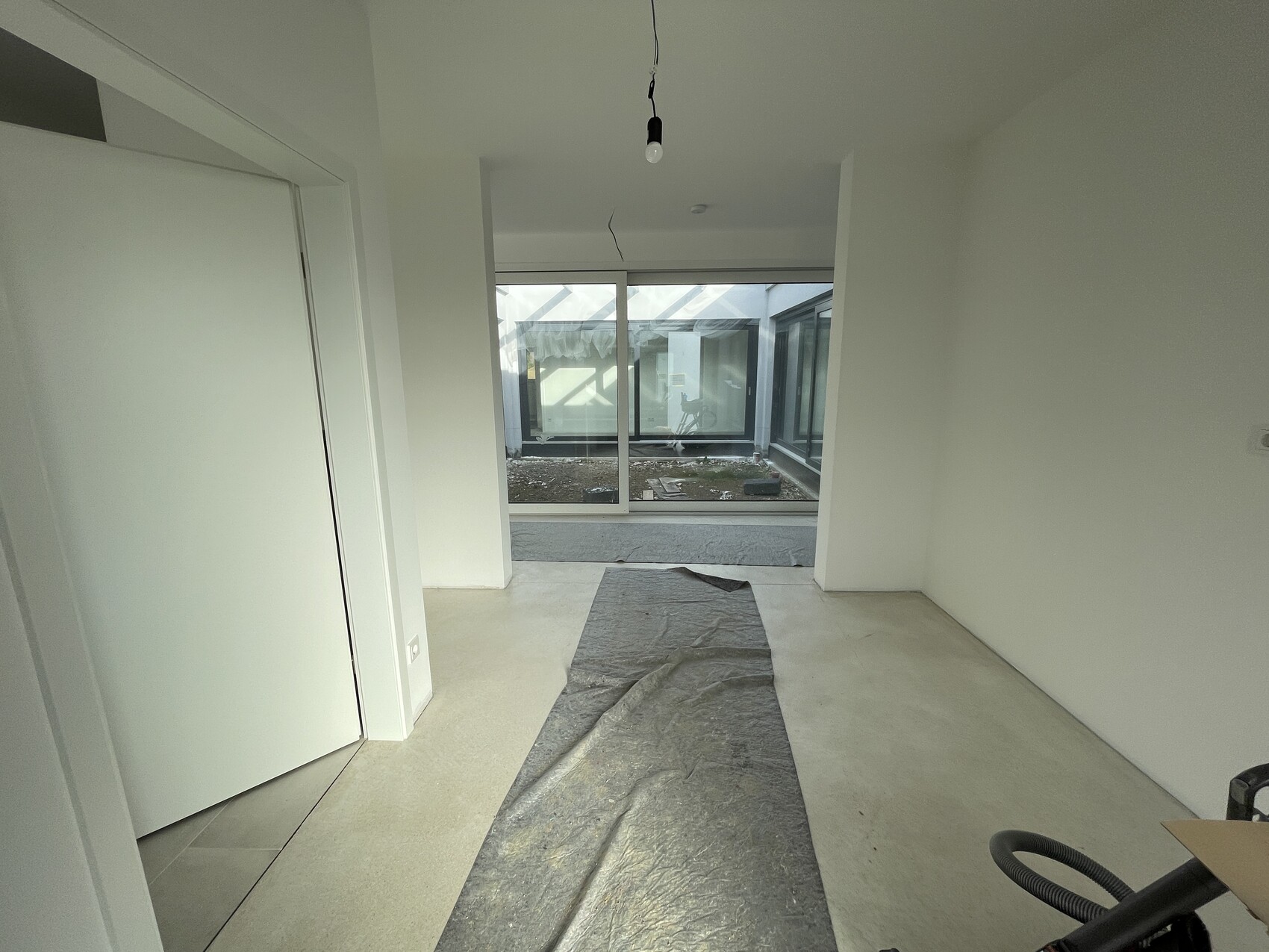 Bright unfinished interior with exposed light bulb, protective floor covering, and glass wall facing the courtyard.