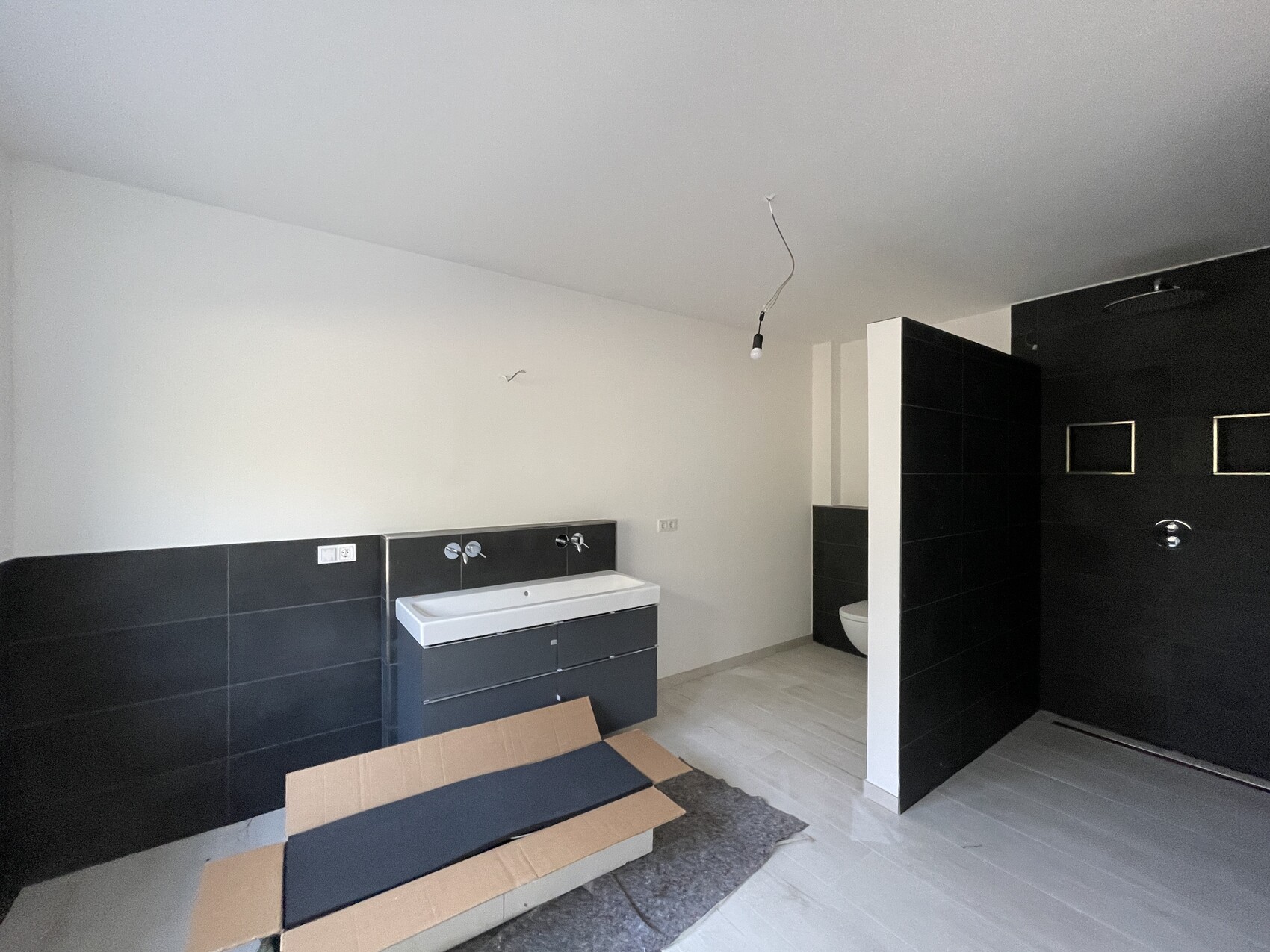 New bathroom with black tiles, washbasin, toilet behind partition wall, exposed cable light.