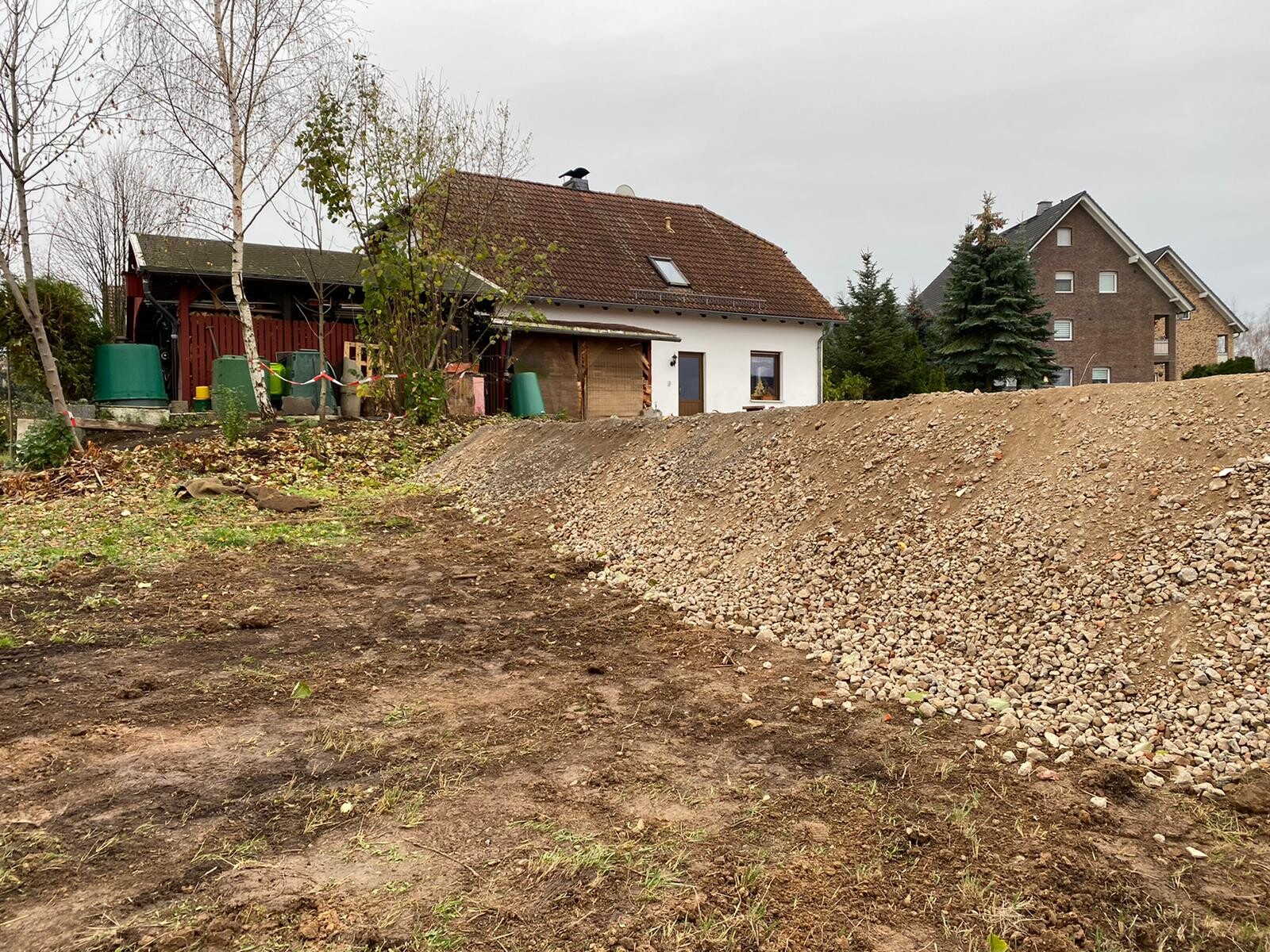 Baustelle: Erdhügel und Kieshaufen neben einem weißen Haus mit Bäumen im Hintergrund.