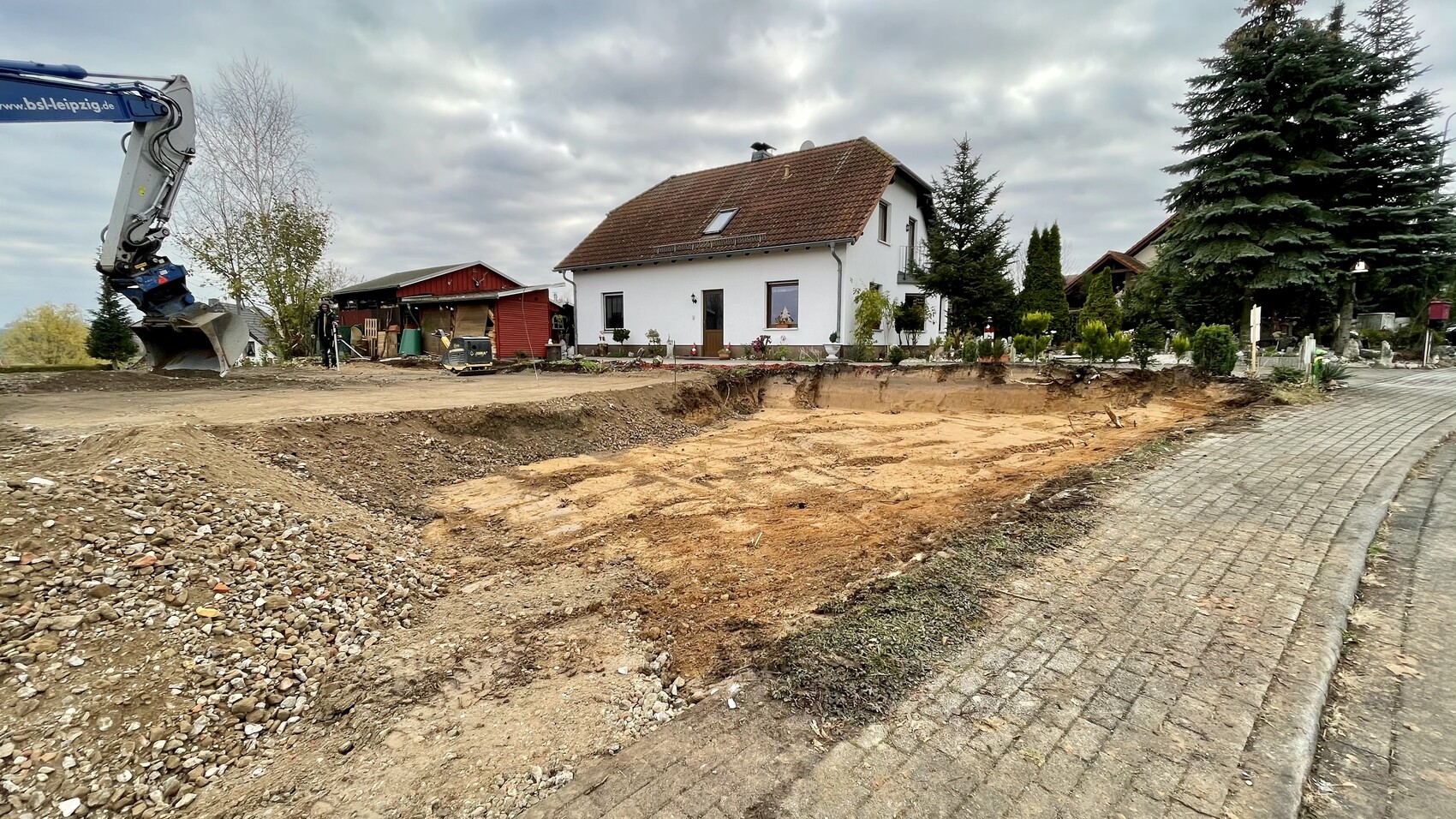 Baugrube vor weißem Einfamilienhaus mit rotem Dach, Baugerät links, Gehweg rechts.