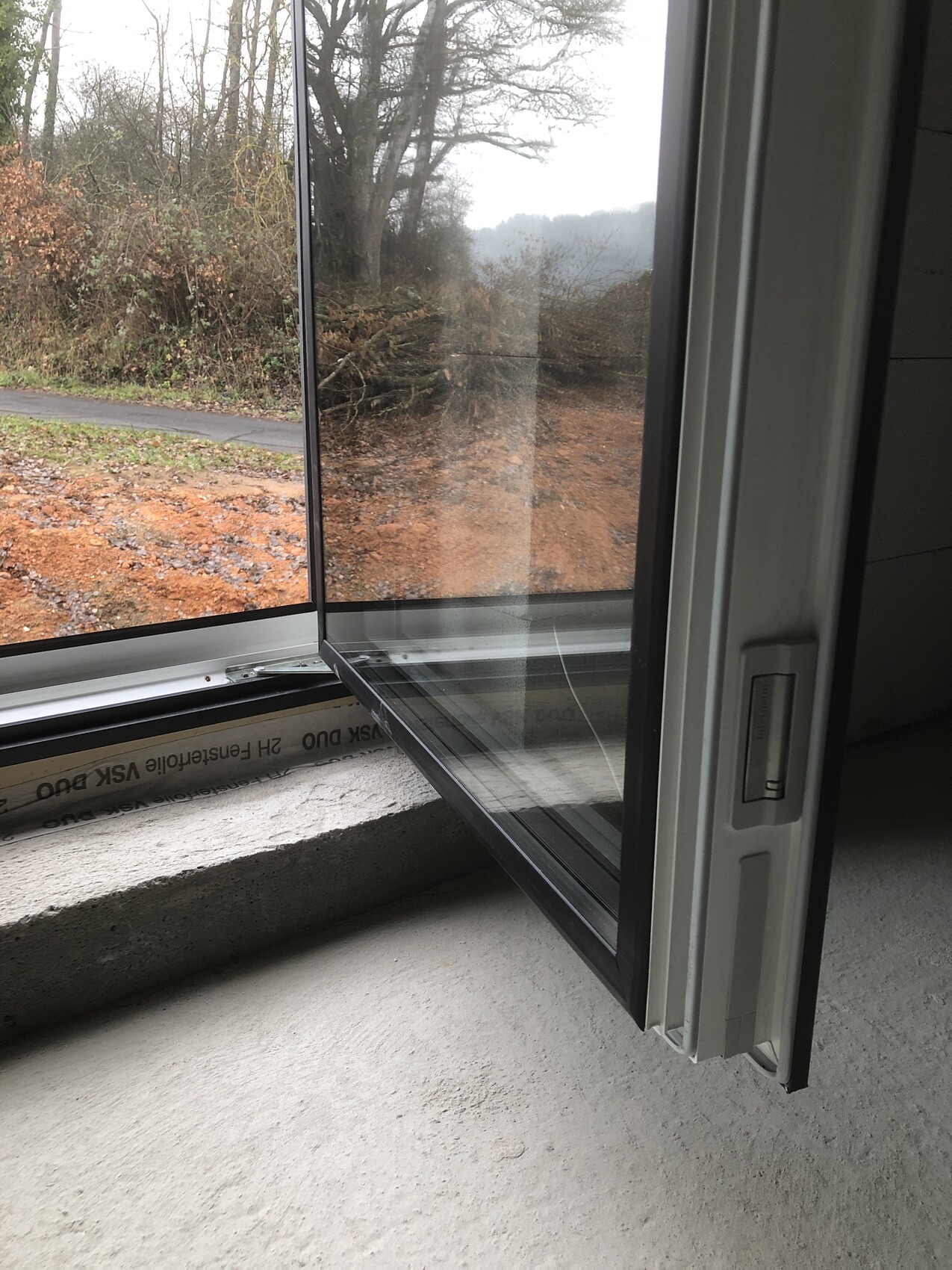 Open window; view outside to path, bushes, and trees; exposed concrete floor visible.