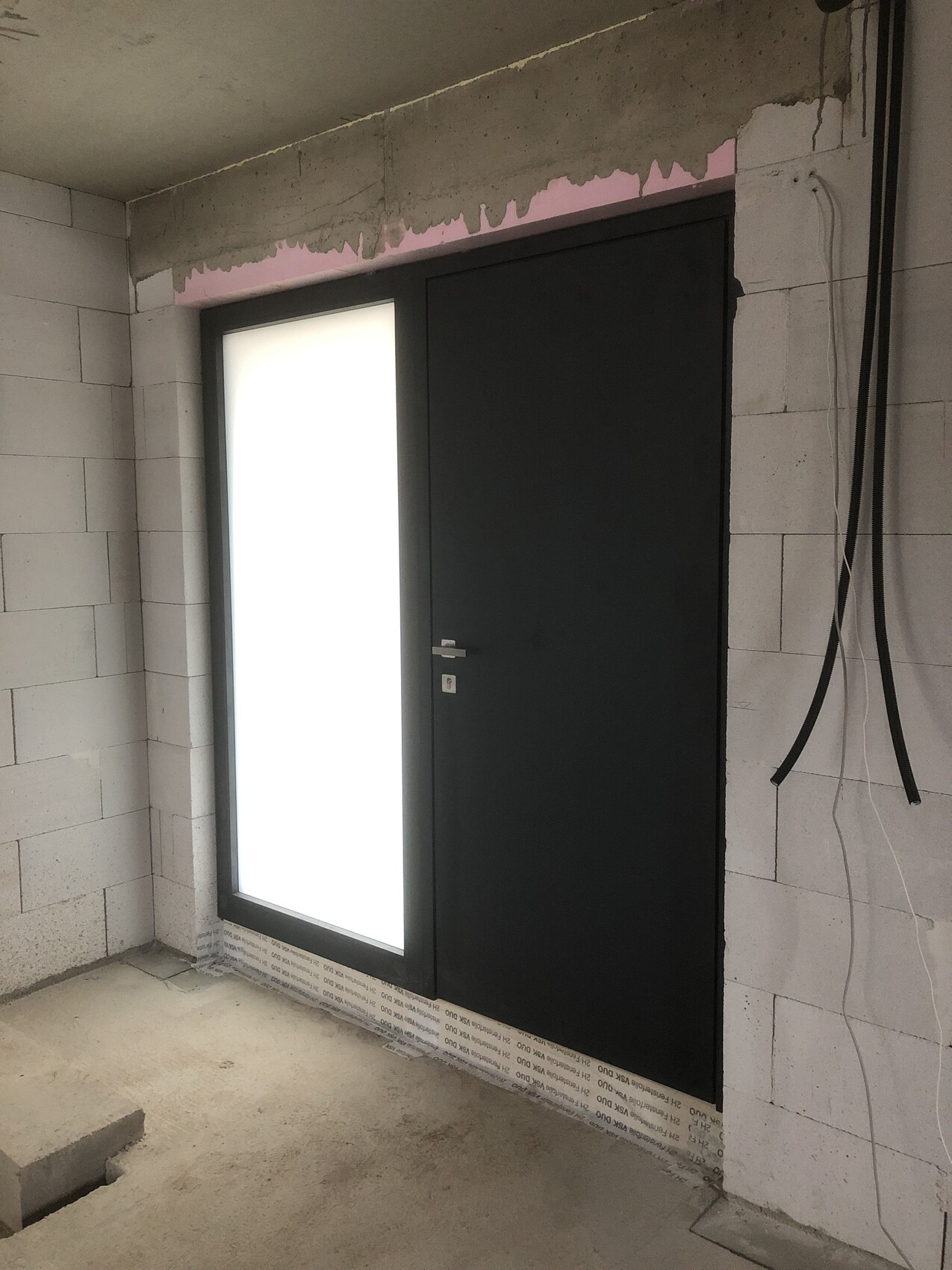 Shell construction: dark door next to milky white glass window; cables on wall, unfinished concrete walls.