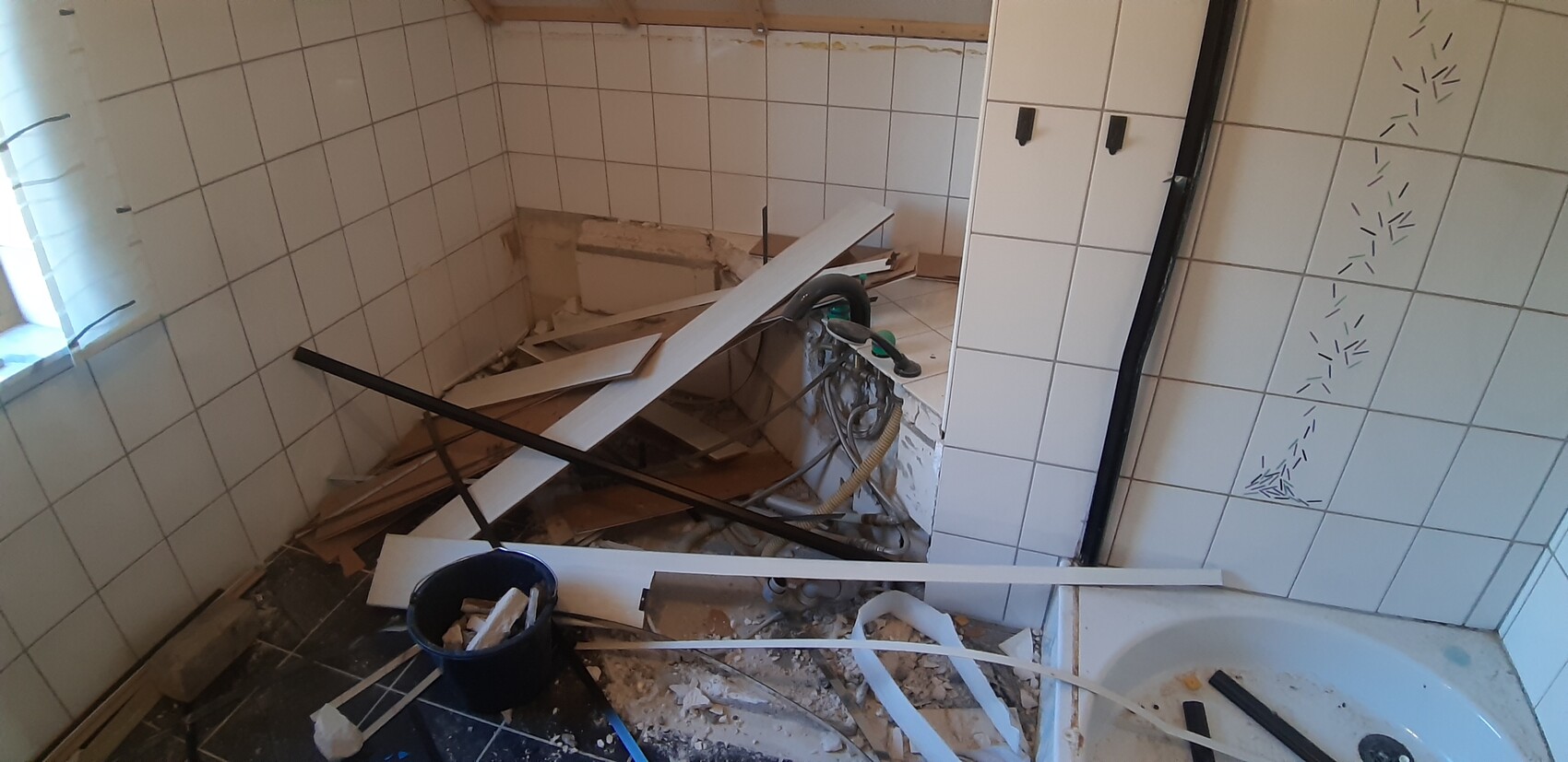 Bathroom under renovation: scattered wooden slats, broken tiles, and exposed pipes around a bathtub.