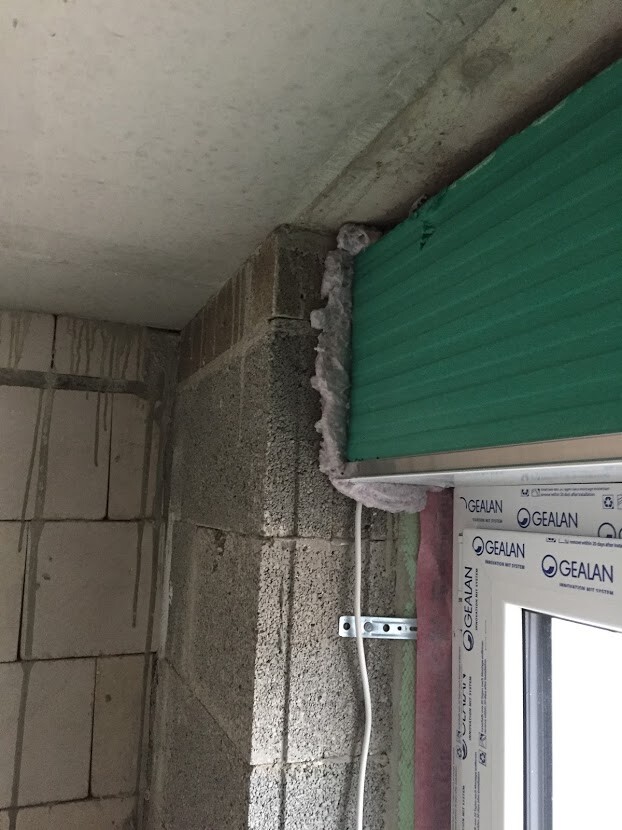 Unfertige Bauwand aus Betonsteinen mit grünem Rollladen, Dichtmasse und Kabel.