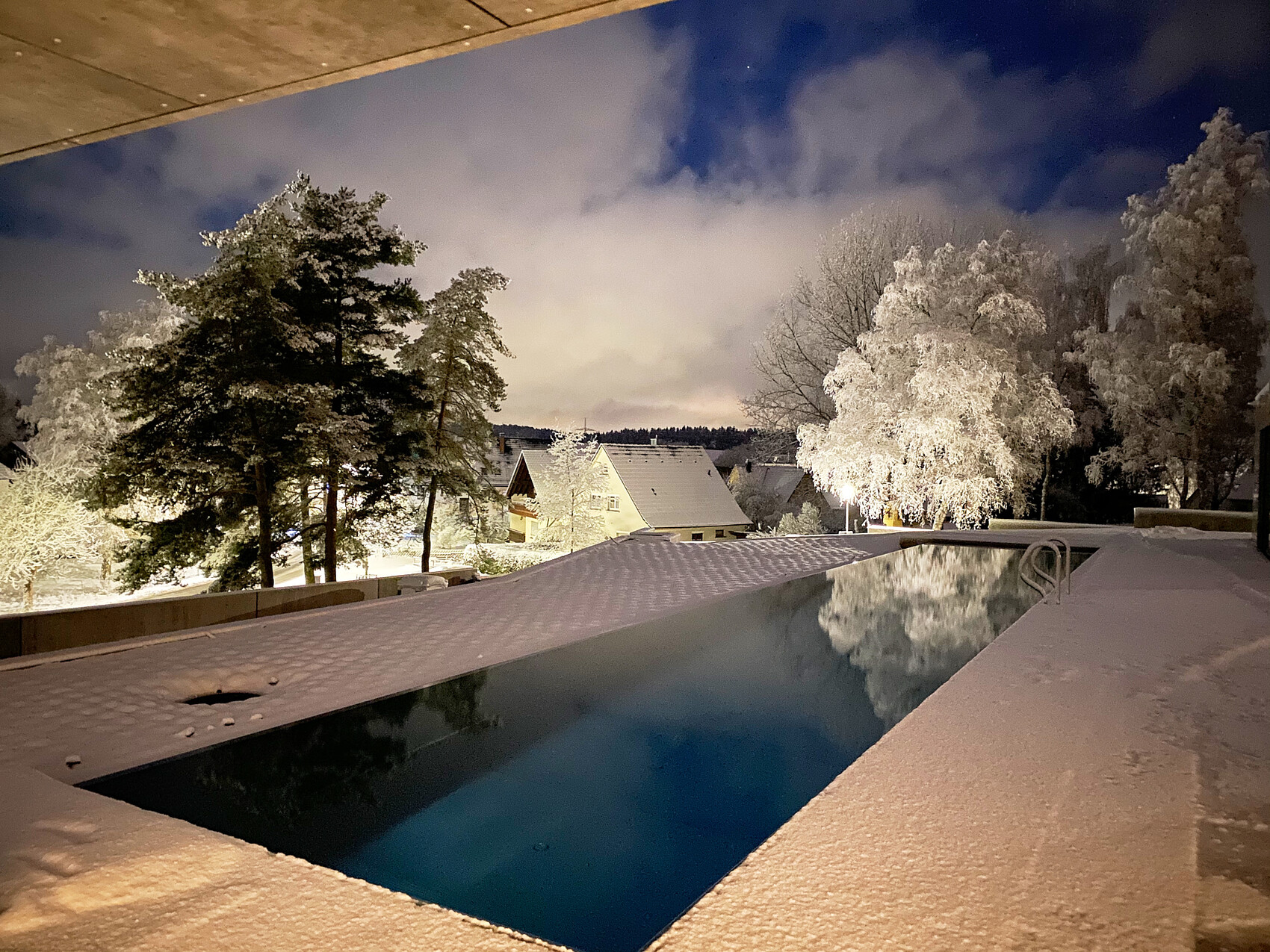Night shot of a snow-covered terrace with pool, trees, and houses in the background.