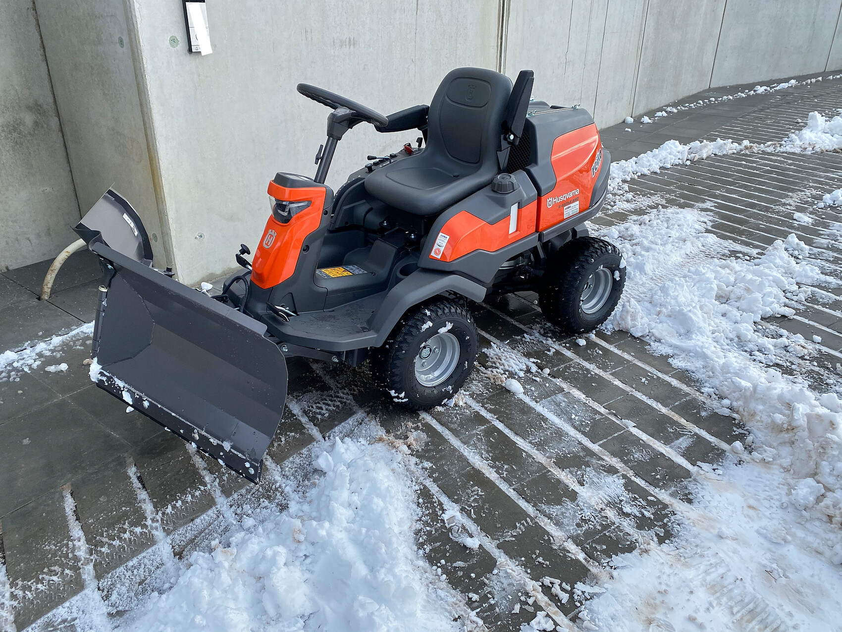 Small orange-gray Husqvarna snowplow with seat and front blade in the snow.