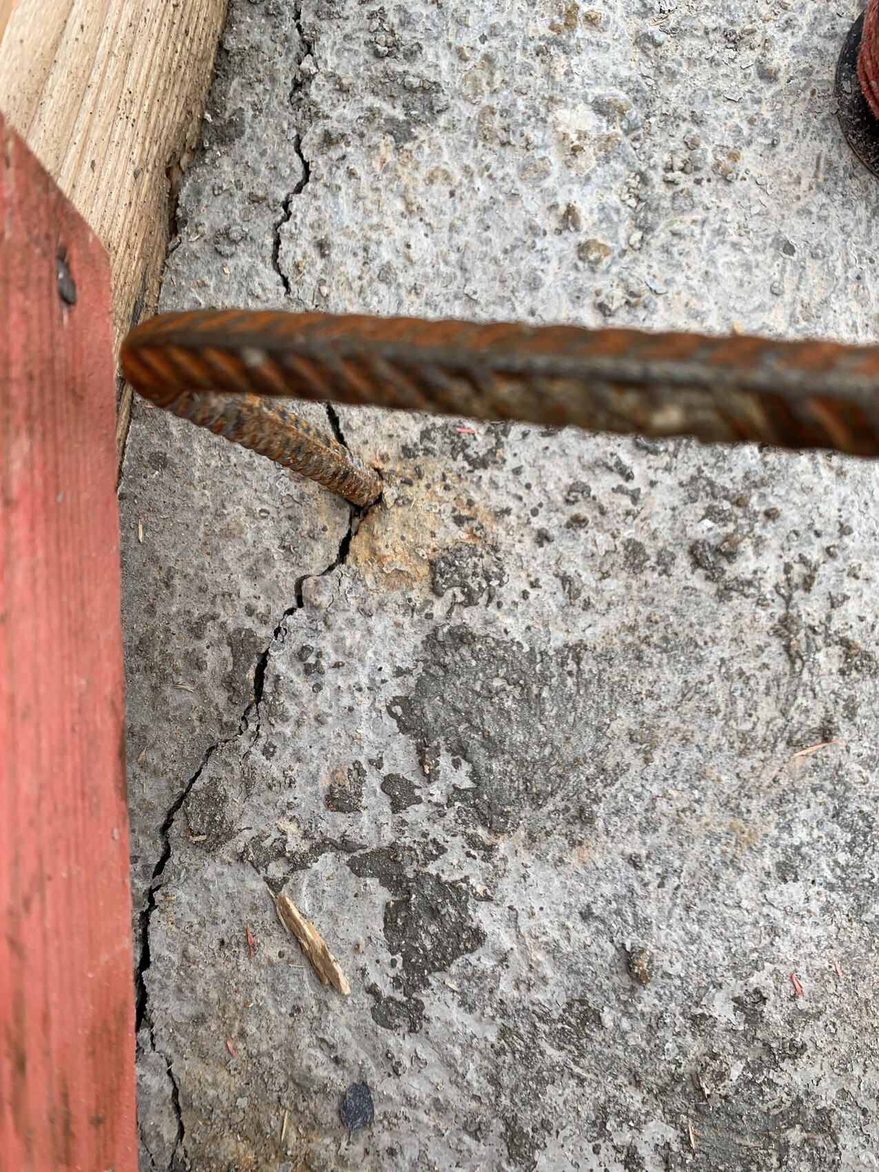 Rusty reinforcing bar protruding from cracked concrete floor, with a red wooden board visible to the left.