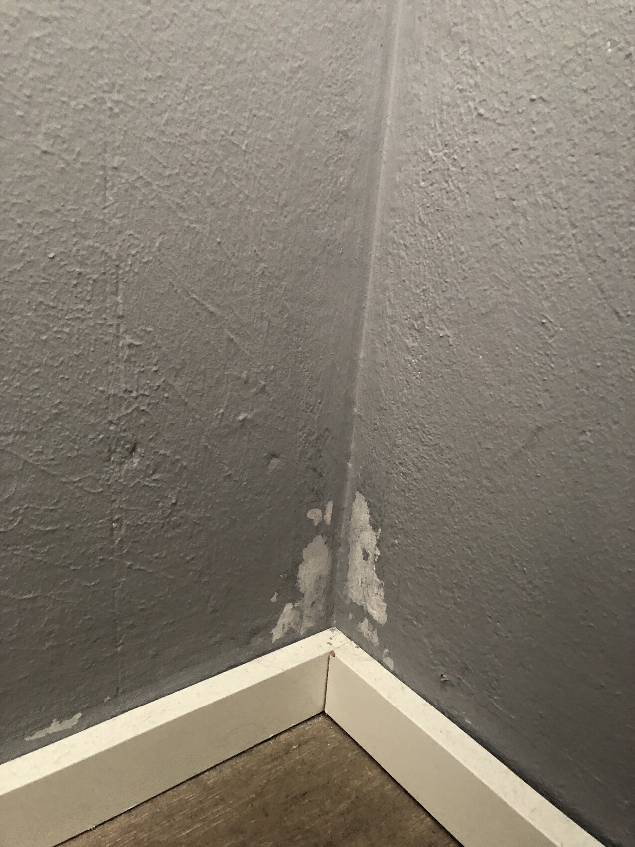 Corner of two gray walls with chipped paint/plaster near the white baseboard.