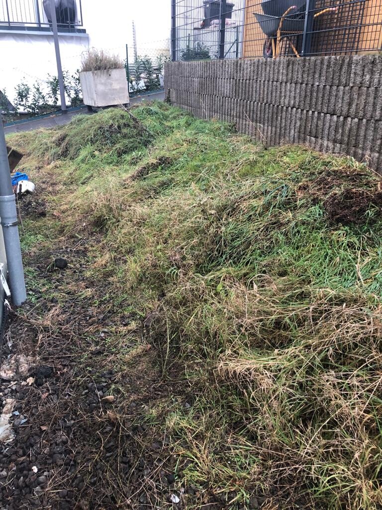 Grasbewachsene Erdaufschüttung neben Gebäudemauer mit Zaun; links sichtbar Abflussrohr.