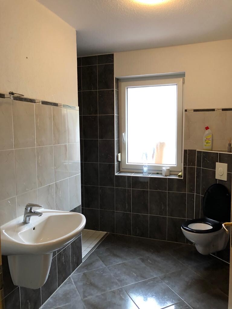 Badezimmer mit Waschbecken links, Toilette rechts, dunkle Fliesen, Fenster mittig.